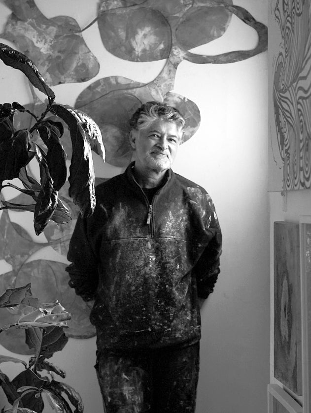 Portait noir et blanc de l'artiste colombien Gustavo Vejarano posant devant un mur orné de ses sculptures. Un artiste abstrait représenté la la Galerie Vermeulen.