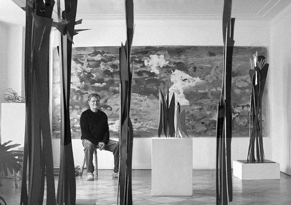 L’artiste Gustavo Vejarano assis dans une salle d’exposition, entouré de ses sculptures verticales et d’une grande toile abstraite en arrière-plan – photographie en noir et blanc présentée par la Galerie Vermeulen.