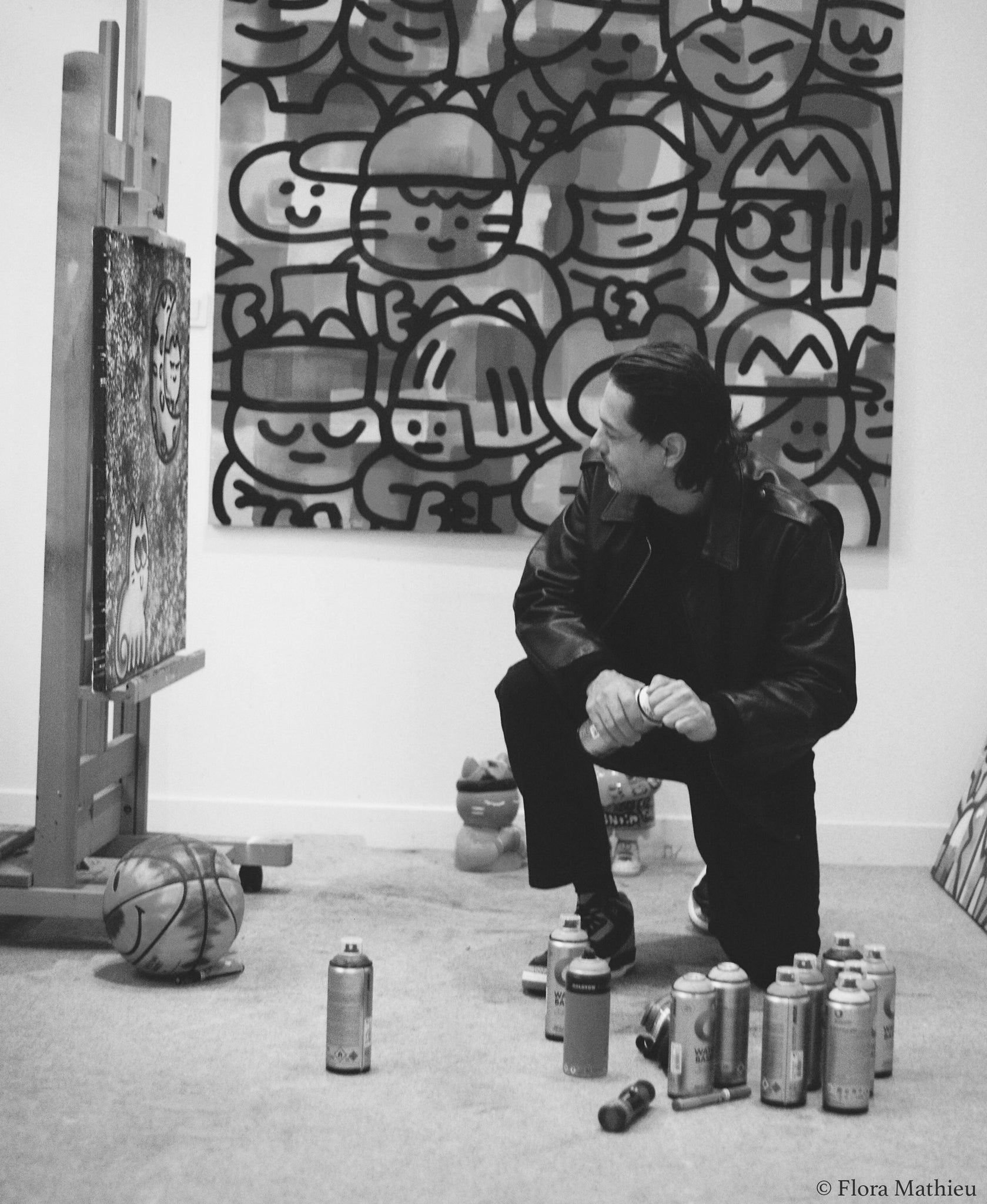 Portrait noir et blanc de l'artiste Chanoir - représentée par la Galerie Vermeulen - dans son atelier parisien. L'artiste un genou au sol, tient une bombe de peinture acrylique dans ses mains et s'apprête à peindre sa toile. Crédit photo : Flora Mathieu.