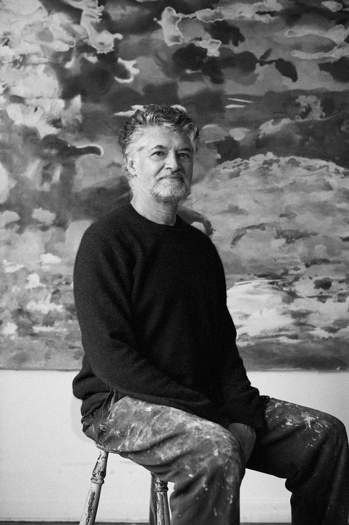 L’artiste Gustavo Vejarano assis dans une salle d’exposition, devant une grande toile abstraite en arrière-plan – photographie en noir et blanc présentée par la Galerie Vermeulen.