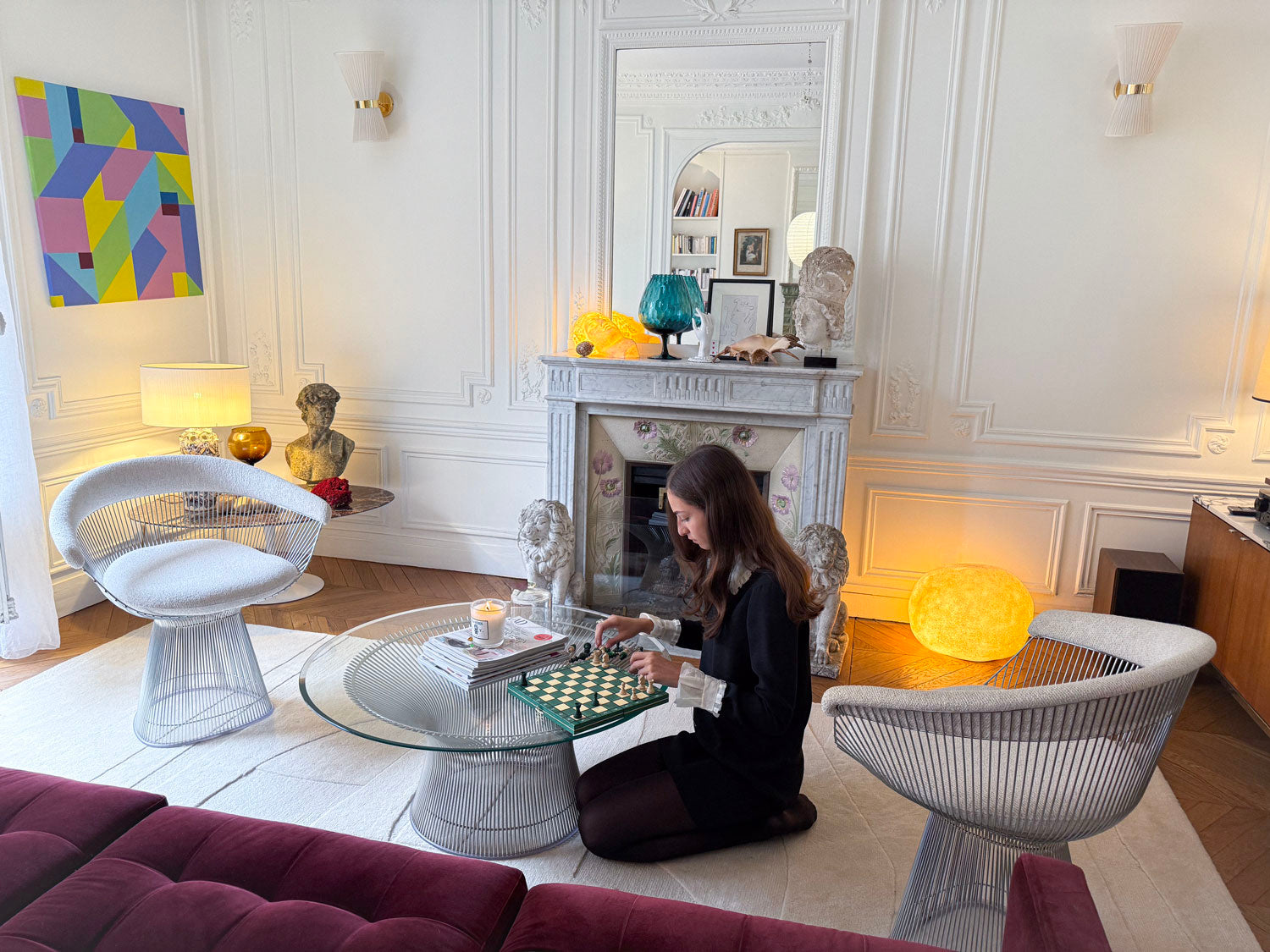 Intérieur haussmannien à Paris, salon design avec chaises Platner, cheminée en marbre, bustes antiques et œuvre contemporaine de Debens ; une collectionneuse joue aux échecs, atmosphère élégante et lumineuse.