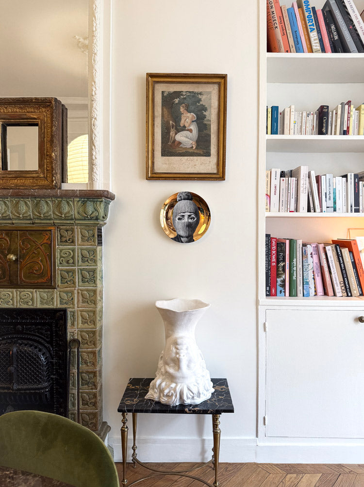 Coin de salon haussmannien avec cheminée en faïence verte, étagères de livres, assiette Fornasetti au mur et vase sculptural blanc sur table en marbre.