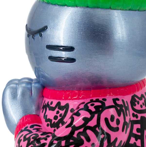Gros plan de profil gauche de l’épreuve d’artiste CHANOIR – Sculpture unique peinte à la main. Chat bleu argenté aux yeux clos, coiffé d’un bonnet vert vif, vêtu d’un pull rose à motifs graffiti noirs. Pièce exclusive fusionnant street-art et pop art et sculpture contemporaine.